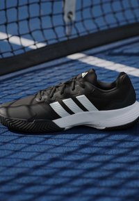 Μαύρο και λευκό παπούτσι τένις Adidas σε μπλε γήπεδο τένις με ένα δίχτυ που ρίχνει σκιά πάνω στο παπούτσι και το γήπεδο.