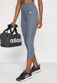 Γκρι αθλητικό κολάν με ψηλή μέση, διακοσμημένο με μια διακριτική ραφή και το λογότυπο της Adidas. Στο χέρι κρατιέται μία μαύρη τσάντα duffel με λευκή επωνυμία Adidas.