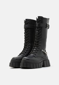 Μαύρες μπότες μέχρι το γόνατο με κορδόνια, κατασκευασμένες από συνθετικό υλικό, που διαθέτουν χοντρή σόλα, φερμουάρ στο πλάι και ρυθμιζόμενο λουράκι με αγκράφα.