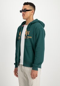 Sudadera verde con cremallera hecha de tejido suave, con un bolsillo tipo canguro, un logotipo naranja y puños acanalados. Lleva puesta sobre una camiseta blanca.