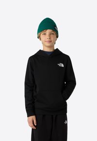 Hanorac negru cu buzunar frontal și manșete striate, cu un mic logo alb. Purtat cu pantaloni de trening negri și o căciulă tricotată verde.