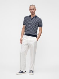 Homme debout en pose décontractée portant un polo bleu marine à rayures manches courtes, un pantalon blanc et des baskets bleu marine sur fond blanc uni.