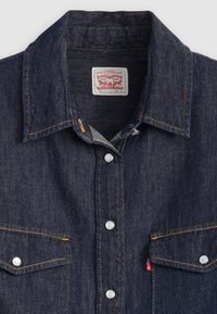 Mörkblå denimskjorta med spetsig krage, två bröstfickor, vita tryckknappar och orange sömmar. Etiketten säger "Levi Strauss & Co."