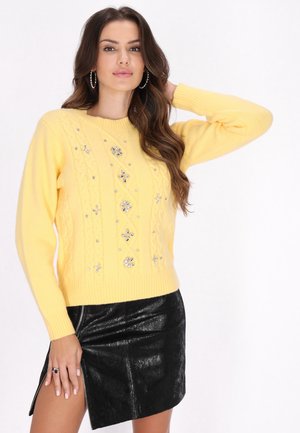 Vrouw met lang bruin haar die een gele trui met decoratieve stenen draagt en een zwarte, getextureerde leren rok met een zijsplit.