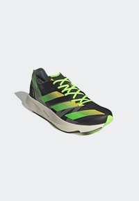 Chaussure de running noire avec des bandes vert fluo et jaunes, des lacets vert fluo et une semelle blanche sur un fond gris clair uni.