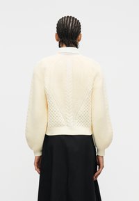 Άτομο με πλεξούδες που φοράει εκρού πουλόβερ από πλεκτό με σχέδιο καλώδιο και φουσκωτά μανίκια και μαύρη φούστα, εμφανιζόμενο από πίσω μπροστά σε λευκό φόντο.