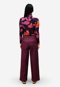 Femme aux longs cheveux noirs portant un haut à manches longues coloré avec un motif et un pantalon bordeaux à larges rayures, se tenant de dos.