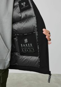 Schwarze Jacke mit einer gesteppten grauen Innenauskleidung und einer Techniktasche. Merkmale sind das Markenlabel, Reißverschlussdetails und Anweisungen auf der Innenauskleidung.