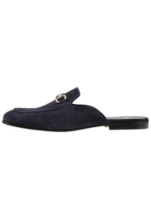 Laivastonsininen mokkainen slip-on-muli hopeisella metallisella hevosenkenkäkoristeella ja mustalla litteällä pohjalla, sivukuva valkoisella taustalla.