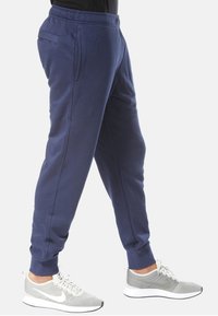 Marinblå mjukisbyxor med elastisk midjeresår, sidofickor och avsmalnande muddar. Kombinerade med ljusgråa sportskor. Slät textur.