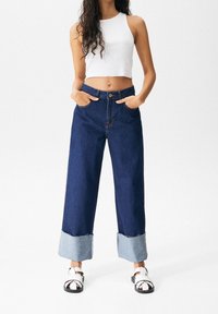 Vaqueros anchos azul con dobladillo sin acabar y puños azul claro, usados con una camiseta de tirantes corta blanca y sandalias blancas de tiras.