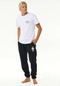 Hvid bomulds T-shirt med sort logo på brystet, parret med sorte sweatpants med logo og farverigt grafik på venstre ben.