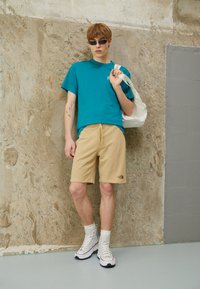 T-shirt azzurro a girocollo, pantaloni shorts beige con logo, sneaker bianche alte e borsa a tracolla in tela, su uno sfondo di muro testurizzato.