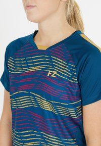 Kvinde iført en blå sports t-shirt med røde og gule bølgemønstre og "FZ" logo på brystet, hår samlet i hestehale.