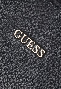 Surface en cuir noir texturé avec des lettres en métal argenté en relief formant le mot « GUESS » en diagonale.