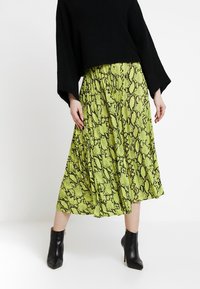 Jupe midi verte à motif serpent avec des plis, faite en tissu léger. Portée avec un pull cropped noir et des bottines noires.