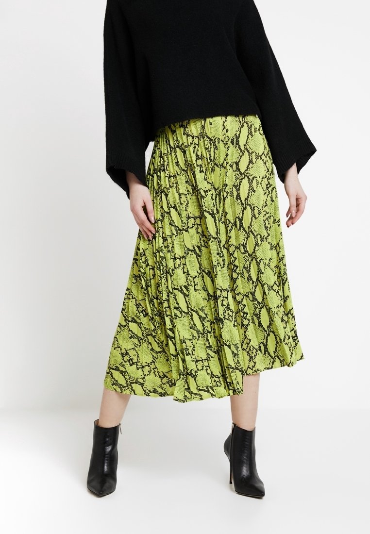 Jupe midi verte à motif serpent avec des plis, faite en tissu léger. Portée avec un pull cropped noir et des bottines noires.