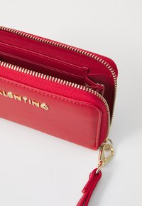 Raudona zip-around piniginė su auksiniais elementais, tekstūruota išorė ir raktų pakabukas. Yra daug kortelių lizdų ir skyrių.