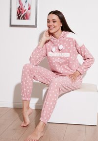 Ružový fleece loungewear set s bielymi bodkami, obsahujúci kapucňu, bambule a štruktúrovaný akcent na pleci. Mäkka a príjemná tkanina.