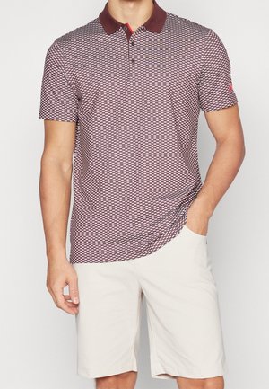 Homme portant un polo à manches courtes à motif bordeaux et un short blanc à hauteur des genoux, avec une main dans la poche.