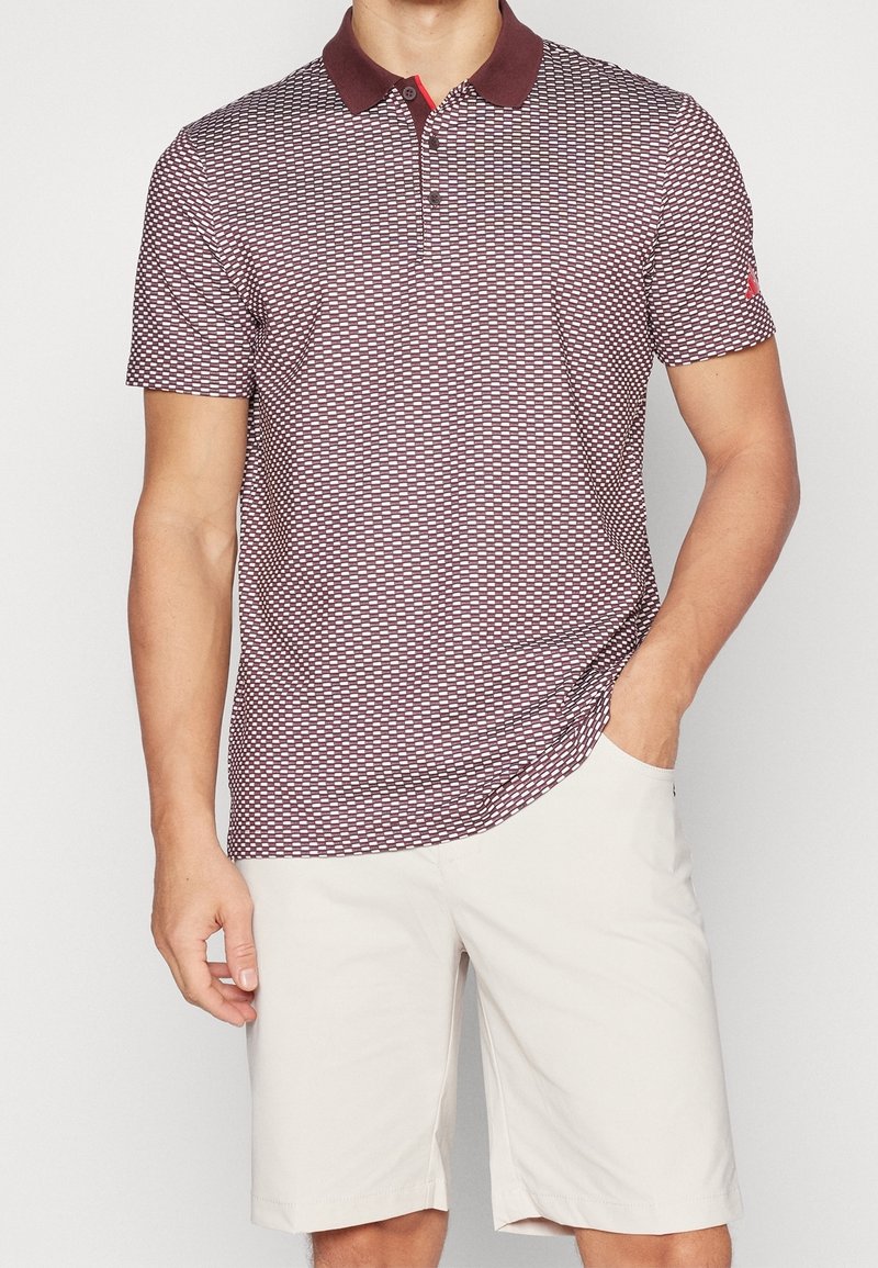 Homme portant un polo à manches courtes à motif bordeaux et un short blanc à hauteur des genoux, avec une main dans la poche.