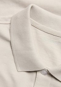 Licht beige polo shirt van een textiel met textuur, ribbelkraag en een plastic knoop aan de voorkant. Etiketten zijn gedeeltelijk zichtbaar.