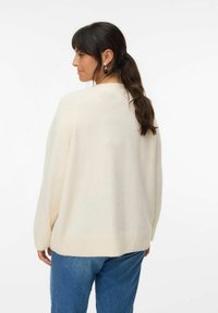 Krémový oversized svetr s ribbovaným lemem, vyrobený z měkké tkaniny, nošený s modrými džíny, viditelné švy a minimální detaily.