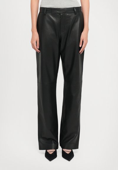 Mujer que lleva pantalones de cuero negro de pierna ancha de cintura alta, combinados con zapatos negros de punta afilada, de pie frente a un fondo blanco liso.