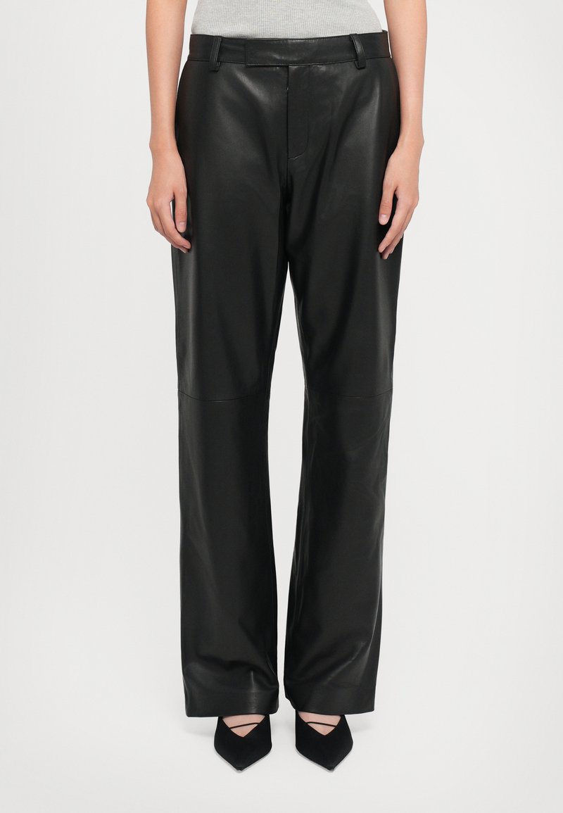 Woman wearing high-waist black leather wide-leg pants paired with black pointed-toe shoes, standing against a plain white background.