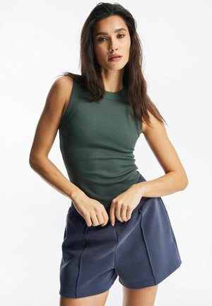 Kobieta o długich ciemnych włosach, ubrana w zielony prążkowany top bez rękawów i granatowe szorty, poprawiająca pasek szortów.
