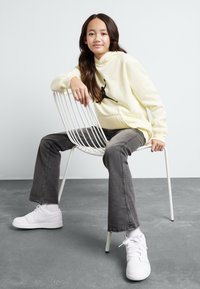Sudadera amarilla con capucha y un logo negro, jeans acampanados gris oscuro y zapatillas blancas, sentado en una silla de alambre blanca contra un fondo claro.
