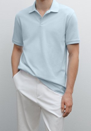 Mand iført lyseblå kortærmet poloshirt og hvide bukser, med den ene hånd i lommen og den anden afslappet ved siden.