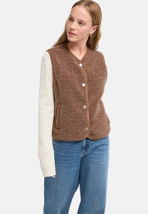 Jeune femme portant une veste polaire marron boutonnée sur une chemise blanche à manches longues et un jean bleu, regardant légèrement vers sa gauche.