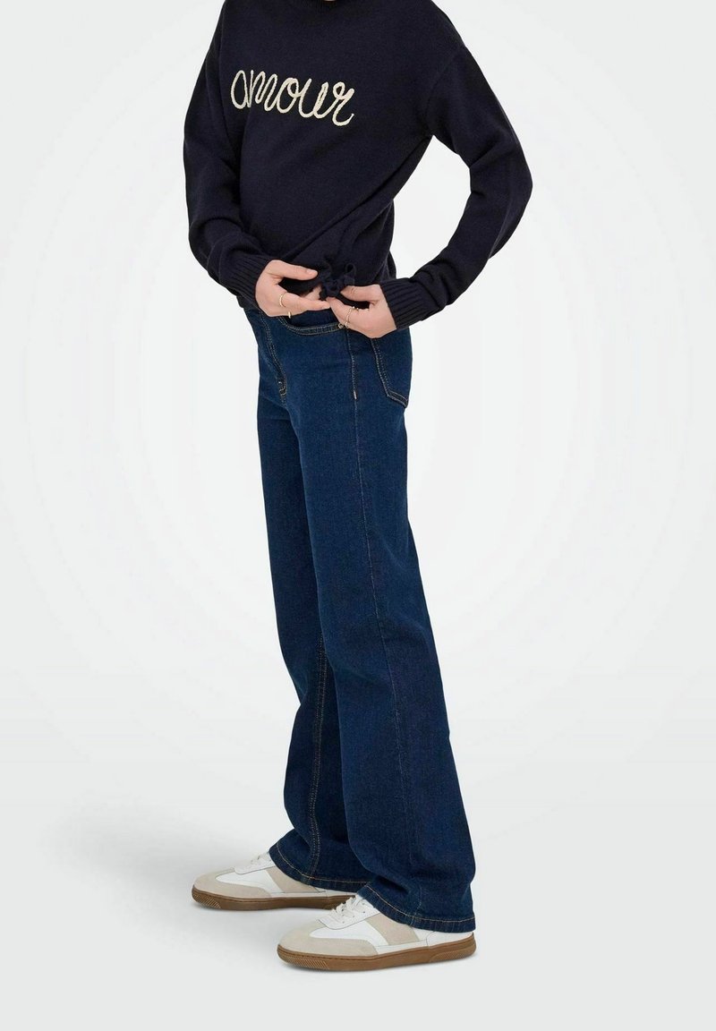Personne portant un pull bleu marine avec le texte « amour », un jean large bleu foncé et des baskets blanches, debout devant un fond uni.