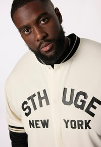 Maglione color crema con scritta nera "STH UGE NEW YORK", dotato di chiusura a bottoni, polsini a righe bianche e nere e un tessuto texturizzato.