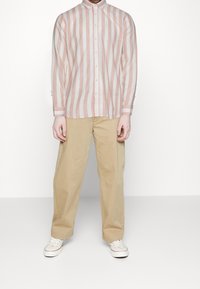 Ljust rosa och vit randig långärmad skjorta med knäppkrage, ihop med beige, löst sittande byxor och vita sneakers.