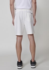 Man som bär vita strukturerade shorts och svarta sneakers, sedd bakifrån mot en enfärgad bakgrund.