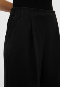 Zwarte geplooide broek met verticale pinstripes, voorzien van zijzakken en een gestructureerde tailleband. De stof lijkt glad en lichtgewicht te zijn.