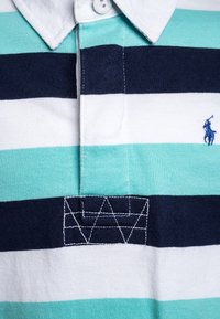 Randig pikétröja i växlande teal, vitt och navy med en logotyp och geometrisk broderidetalj nära kragen.