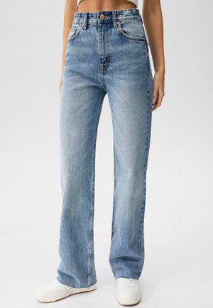 Femme portant un jean droit taille haute bleu clair et des baskets blanches, debout devant un fond clair uni.