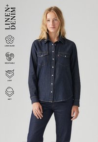 Camisa de denim em mistura de linho azul-escuro com frente abotoada, dois bolsos no peito e botões brancos. Textura suave, design respirável e leve.
