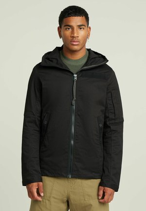 Veste à capuche noire avec fermeture éclair sur le devant, poches latérales et poche sur le bras ; fabriquée en tissu texturé ; présente un col montant et des coutures renforcées.