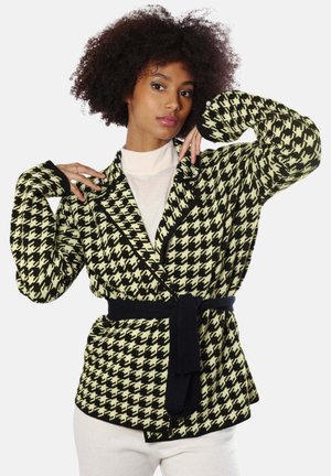 Femme aux cheveux bouclés portant un cardigan ceinturé à motif pied-de-poule noir et blanc, posant avec les mains près du visage et du cou.