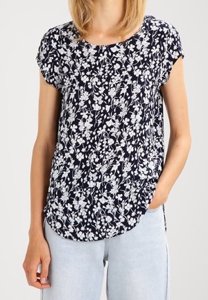 Frau trägt eine marineblaue Kurzarmbluse mit weißem Blumenmuster und hellblauen Jeans, eine Hand in der Tasche, vor einfarbigem Hintergrund.