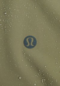 Tessuto verde oliva con gocce d'acqua; un logo blu scuro ben visibile al centro. Texture liscia con una leggera lucentezza.