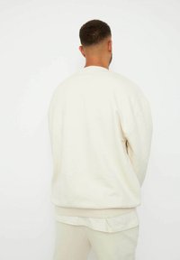 Sudadera de hombre en crema con un corte relajado, puños y dobladillo acanalados. Textura suave, diseño sencillo. Combinada con pantalones claros a juego.