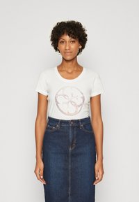 Camiseta blanca de manga corta con un diseño de logo circular de purpurina en el frente, combinada con una falda lápiz de mezclilla de talle alto.