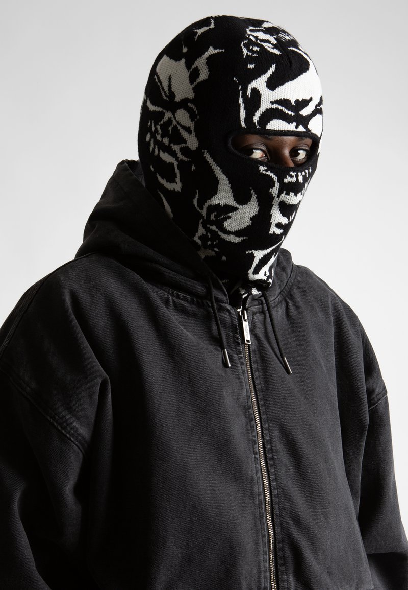 Crna dukserica s patentnim zatvaračem i vezicama, u kombinaciji s crno-bijelim pletenim balaklavom s apstraktnim uzorcima i otvore za oči.