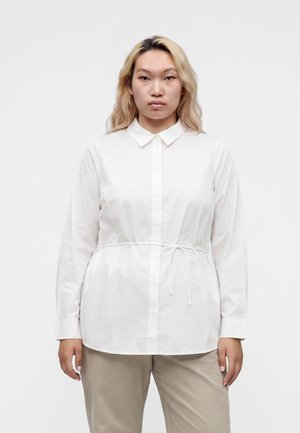 Femme aux longs cheveux blonds, debout face à l’avant, portant une chemise blanche boutonnée avec ceinture à la taille et un pantalon beige sur un fond uni.