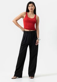 Top aderente rosso abbinato a pantaloni neri a vita alta con pieghe. Il modello indossa sandali neri con cinturini e un bracciale trasparente al polso.